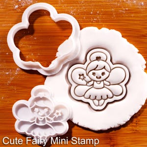 May include: A white plastic cookie cutter and a matching stamp featuring a fairy design with wings and a star wand. The stamp is pressed into a circular piece of white dough, showing the fairy outline. The text Cute Fairy Mini Stamp is visible.