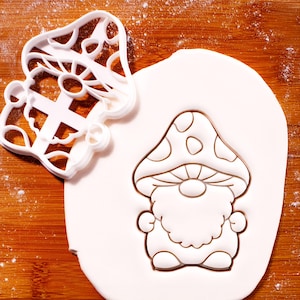 May include: A white plastic mushroom-shaped cookie cutter next to a fondant cutout of a gnome with a mushroom cap. The gnome has a beard and is wearing a mushroom cap with spots. The background is a wooden surface.