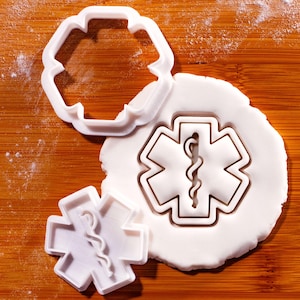 Może przedstawiać: Biała plastikowa foremka do ciastek i wytłoczone ciastko z symbolem Gwiazdy Życia, sześcioramienną gwiazdą z laską i wężem. Foremka leży na drewnianej powierzchni.