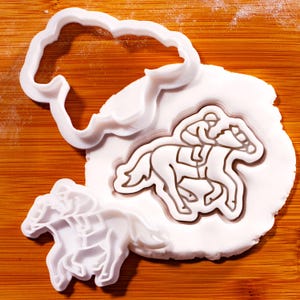Op de afbeelding: Witte plastic koekjessnijders in de vorm van een paard en jockey, en een wolk. Het paard en de jockey zijn in een wit deeg gedrukt. De uitstekers liggen op een houten oppervlak.