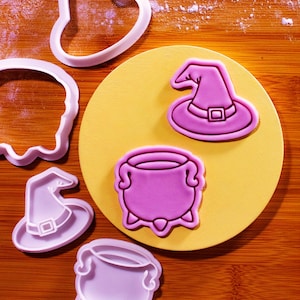 Op de afbeelding: Een verzameling roze plastic Halloween-koekjessnijders. De uitstekers bevatten een heksenhoed, een ketel en andere vormen. De uitstekers zijn gerangschikt op een gele cirkel en een houten oppervlak.