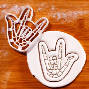 Può includere: Un tagliapasta in plastica bianca a forma di mano scheletrica che fa il segno "Ti amo", accanto a un'impronta di fondente bianco dello stesso disegno. Il tagliapasta è su una superficie di legno con una spolverata di polvere bianca.