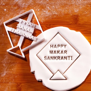 May include: A white plastic kite-shaped cookie cutter next to a white fondant cutout. The fondant has the words "HAPPY MAKAR SANKRANTI" embossed within a kite shape. The cutter has the same text.