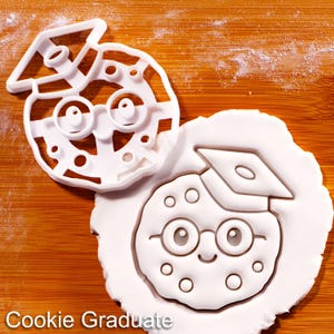 May include: A white plastic cookie cutter and a cookie with an embossed design. The cutter is in the shape of a graduate cookie with a mortarboard hat and glasses. The cookie has a smiling face and is on a wooden surface. The text "Cookie Graduate" is at the bottom.