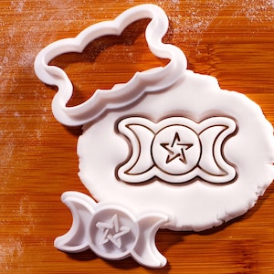 May include: White plastic cookie cutters shaped like a cloud and a triple moon symbol with a star. The cutters are on a wooden surface with white dough. The triple moon symbol is also imprinted in the dough.