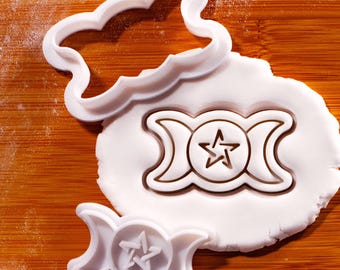 May include: White plastic cookie cutters in the shape of a cloud and a triple moon symbol with a star. The cutters are on a wooden surface with white dough. The triple moon symbol is also imprinted in the dough.