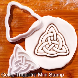 May include: White plastic Celtic Triquetra mini stamp and matching cookie cutter on a wooden surface. The stamp is pressed into white dough, creating a detailed design. The text "Celtic Triquetra Mini Stamp" is at the bottom.