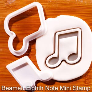Op de afbeelding: Een witte plastic koekjesvorm in de vorm van een achtste noot, een bijpassende witte plastic stempel en een cirkelvormig stuk wit deeg bedrukt met het achtste noot ontwerp. De tekst Beamed Eighth Note Mini Stamp is zichtbaar.