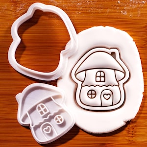 May include: Two white plastic cookie cutters shaped like a mushroom house with a rounded roof, a chimney, two windows, and a heart-shaped door. One cutter is pressed into a circular piece of white dough, showing the embossed house design.