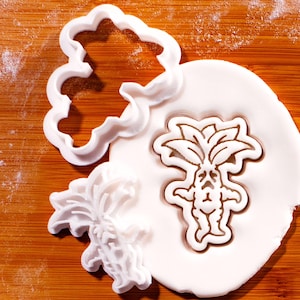 May include: White plastic cookie cutters in various shapes, including a cloud and a mandrake. One mandrake shape is pressed into white dough, showing a detailed outline. The cutters are on a wooden surface.