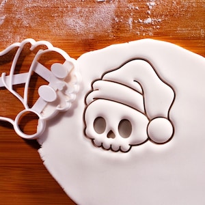 May include: A white skull-shaped cookie cutter with a Santa hat design next to a cut-out skull shape in white dough. The cutter has a detailed design and is on a wooden surface.