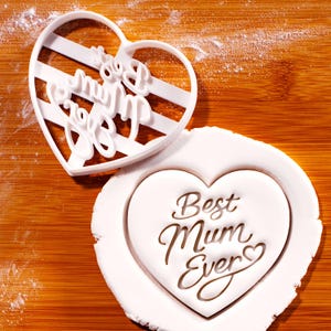 May include: A white heart-shaped cookie cutter with the words "Best Mum Ever" and a matching cookie. The cutter has a striped design. The cookie is white with the words "Best Mum Ever" imprinted on it. The image is on a wooden surface.