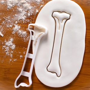 May include: White plastic cookie cutter shaped like a bone. The cutter is on top of a white fondant surface.