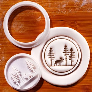 Könnte beinhalten: Weiße Ausstecher aus Kunststoff mit einem Wald- und Elch-Design. Das Set enthält einen runden Ausstecher, einen kreisförmigen Stempel mit einem Elch und Bäumen sowie einen kleineren Stempel mit ähnlichem Design. Geeignet für Kekse.