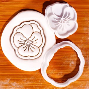 以下が含まれることがあります： 木製の表面に置かれた、3つの白いプラスチック製の花型のクッキーカッター。1つは完全な花の形、もう1つは詳細な花、3つ目は花の波状の輪郭です。
