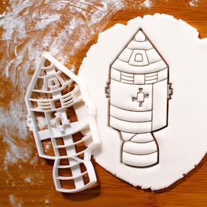 May include: A white plastic cookie cutter in the shape of a rocket ship. The cutter is on a wooden surface covered in white flour.