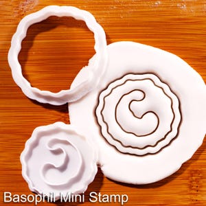 May include: A white plastic cookie cutter with a wavy edge and a matching white plastic stamp featuring a curved, abstract shape. The stamp is shown both as a separate tool and pressed into a piece of white dough, with the text Basophil Mini Stamp.