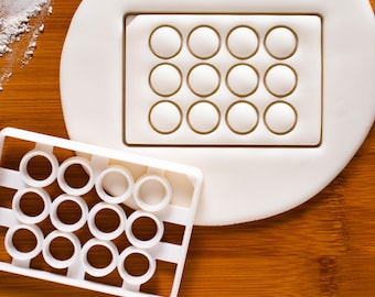 May include: A white plastic cookie cutter with a rectangular frame and twelve circular cutouts. Next to it is a white mold with the same circular pattern. The items are on a wooden surface, with a small amount of white powder in the upper left corner.
