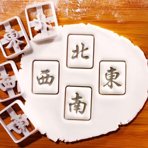 May include: White plastic cookie cutters and stamped cookies with Chinese characters for a Mahjong game. The cookies are stamped with the characters for East, South, West, and North. The cutters are on a wooden surface.