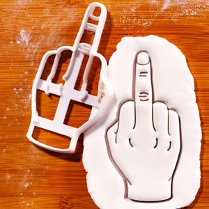 May include: A white plastic cookie cutter in the shape of a hand giving the middle finger, next to a cutout of the same design in white dough. The cutter is on a wooden surface.