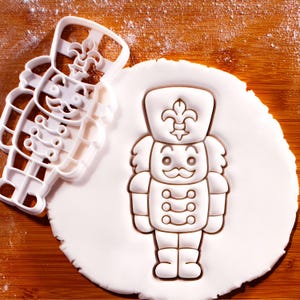 May include: A white plastic cookie cutter and a nutcracker-shaped cookie on a wooden surface. The cookie cutter is in the shape of a toy soldier with a hat and buttons. The cookie is pressed into a white dough.