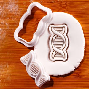 Könnte beinhalten: Ein weißes Ausstechersatz aus Kunststoff mit einem Doppelhelix-Design der DNA. Das Set enthält einen Ausstecher in Form des DNA-Symbols und einen wolkenförmigen Ausstecher. Die Ausstecher befinden sich auf einer weißen Oberfläche mit Holzgrund.