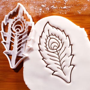 Könnte beinhalten: Ein weißer Ausstecher in Form einer Pfauenfeder neben einem Pfauenfederabdruck auf weißem Teig. Der Ausstecher und der Abdruck zeigen das detaillierte Design der Feder, ideal zum Backen.