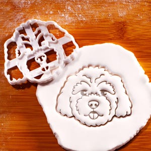 May include: A white plastic dog-shaped cookie cutter next to a white dough cutout of a dog's face. The cutter has a detailed outline of the dog's face, including eyes, nose, and mouth. The dough cutout shows the same dog face design.