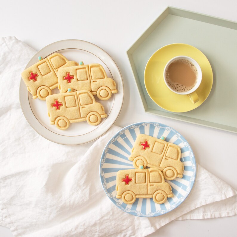 May include: Three yellow ambulance cookies decorated with red crosses on white plates. Two cookies are on a white plate with a gold rim, and one cookie is on a blue and white striped plate. A yellow saucer with a cup of coffee is on a green tray.