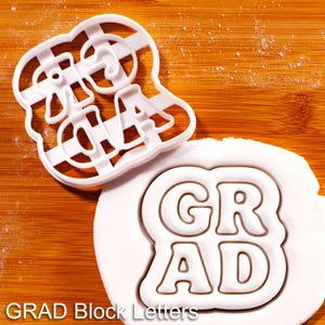 May include: White plastic cookie cutter in the shape of the word "GRAD" in block letters. The letters are cut out, and the cutter has a grid pattern. A white fondant with the "GRAD" letters is next to the cutter. The text "GRAD Block Letters" is at the bottom.