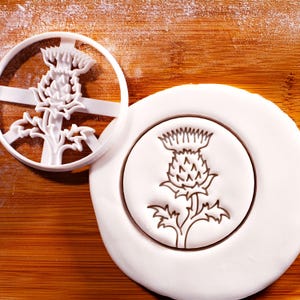 以下が含まれることがあります： 白いプラスチック製のアザミ型クッキーカッターと、アザミのデザインが施された白いフォンダンクッキー。アザミのデザインは茶色で、白いフォンダンに押し込まれています。背景は木製の表面です。