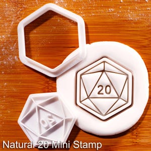 以下が含まれることがあります： クラフト用の白いプラスチック製Natural 20 Mini Stampセット。セットには、サイコロ型のスタンプ、サイコロ型のカッター、六角形カッターが含まれています。スタンプには、幾何学的な形状の中に数字の20が表示されています。