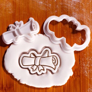 May include: White plastic cookie cutters in the shape of a diploma scroll and a cloud. The scroll cutter is next to a cloud-shaped cutter and a scroll imprint in white dough. The background is a wooden cutting board.