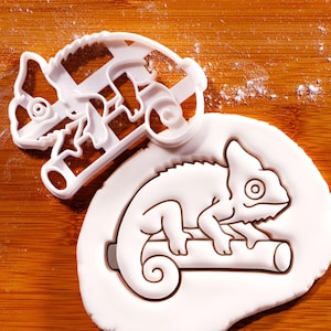 May include: A white plastic chameleon cookie cutter featuring a detailed outline of a chameleon perched on a branch. The cutter is shown alongside a piece of white dough that has been pressed with the cutter to reveal the chameleon shape.