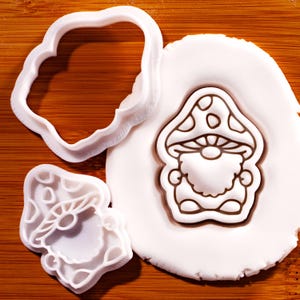 May include: A white plastic cookie cutter shaped like a gnome wearing a mushroom cap, alongside a matching white plastic frame cutter. A piece of white rolled dough shows the embossed outline of the gnome character with a spotted mushroom hat.