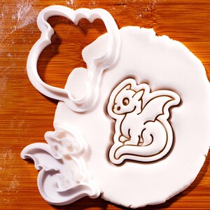 Könnte beinhalten: Ein weißer Drachen-Ausstecher neben einem weißen Fondant-Ausschnitt eines Drachen. Der Ausstecher ist aus Kunststoff und hat einen gebogenen Griff. Der Fondant-Drache hat detaillierte Flügel und ein lächelndes Gesicht. Der Hintergrund ist eine Holzoberfläche.