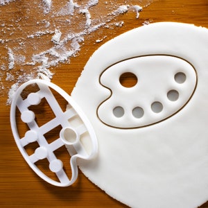 Può includere: Un tagliabiscotti in plastica bianca a forma di tavolozza da pittore accanto a una forma di tavolozza ritagliata in pasta bianca. La farina è cosparsa sulla superficie di legno. Il tagliabiscotti ha un manico e sei rientranze circolari.