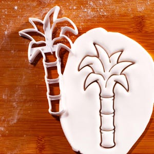 May include: A white plastic palm tree cookie cutter next to a palm tree imprint in white dough. The cutter is a detailed outline of a palm tree, with a segmented trunk and fronds. The dough imprint shows the same design.