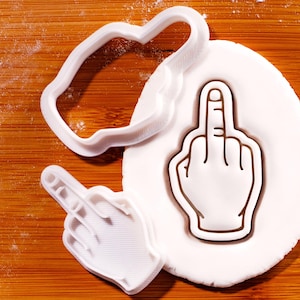 Op de afbeelding: Witte plastic koekjessnijders in de vorm van een hand die de middelvinger opsteekt, met een handvormig koekje. De uitstekers liggen op een houten oppervlak met wit deeg.