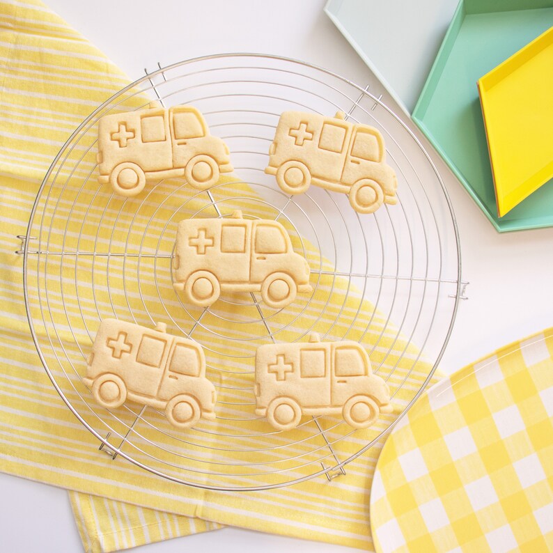 May include: Five yellow sugar cookies shaped like ambulances on a wire cooling rack. The cookies are decorated with red crosses and have a white background.
