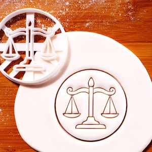 Può includere: Un tagliabiscotti in plastica bianca a forma di bilancia, accanto a un fondente bianco con un disegno di bilancia marrone. Lo sfondo è una superficie di legno con farina.