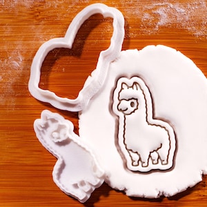 May include: White plastic cookie cutters in the shapes of a heart and an alpaca, along with an alpaca-shaped cookie on a white surface. The cutters are on a wooden cutting board.