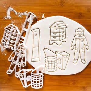 May include: A set of white plastic cookie cutters shaped like a beekeeper, a beehive, a smoker, a ladder, and a honey dipper. The cutters are on a white surface covered in flour.