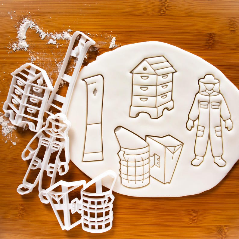 May include: A set of white plastic cookie cutters shaped like a beekeeper, a beehive, a smoker, a ladder, and a honey dipper. The cutters are on a white surface covered in flour.