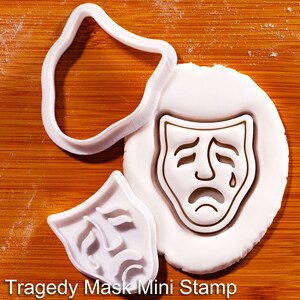 May include: A white plastic cookie cutter and a matching stamp featuring a tragedy mask design. The stamp is pressed into a circular piece of white dough, creating a clear impression of a sad face with a single tear. The text Tragedy Mask Mini Stamp is visible.