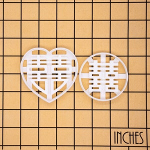 May include: Two white plastic cookie cutters. One is heart-shaped, the other is round. Both feature a Chinese double happiness symbol. The word "INCHES" is printed in the bottom right corner.