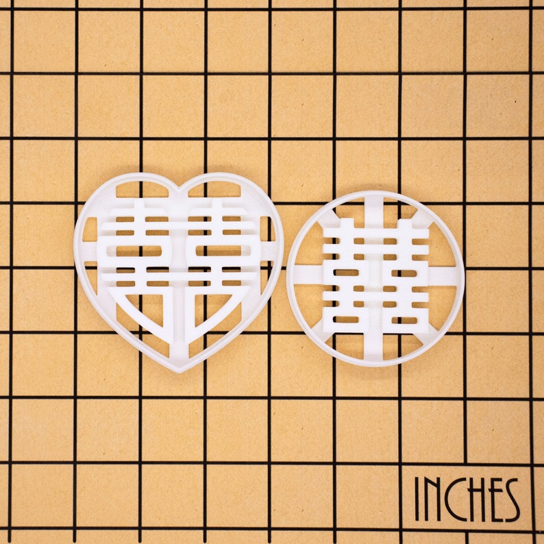 May include: Two white plastic cookie cutters. One is heart-shaped, the other is round. Both feature a Chinese double happiness symbol. The word "INCHES" is printed in the bottom right corner.
