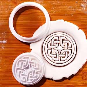 May include: A white plastic biscuit cutter set comprising a circular outer frame and a separate circular stamp with an intricate, symmetrical Celtic knot design. One stamp is shown pressed into a circular piece of white dough, creating a clear impression.