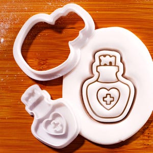 Op de afbeelding: Witte plastic koekjessnijders in de vorm van een hart en een drankfles met een hart en kruis. De drankfles is in witte deeg gedrukt. De achtergrond is een houten oppervlak.