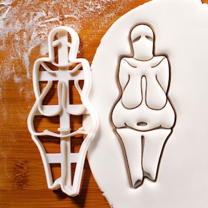 Puede incluir: Dos cortadores de galletas de plástico blanco en forma de figura femenina. El cortador de la izquierda es un contorno detallado del cuerpo de una mujer. El cortador de la derecha es un contorno más simplificado del cuerpo de una mujer.